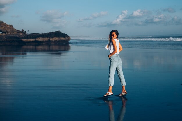 Vrij Kaukasische geschikte vrouw in witte bovenkant en jeans bij het weerspiegelen van strand door oceaan bij zonsonderganglicht