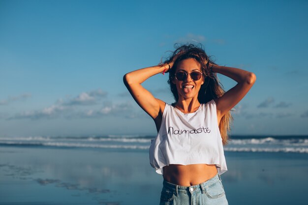 Vrij Kaukasische geschikte vrouw in witte bovenkant en jeans bij het weerspiegelen van strand door oceaan bij zonsonderganglicht