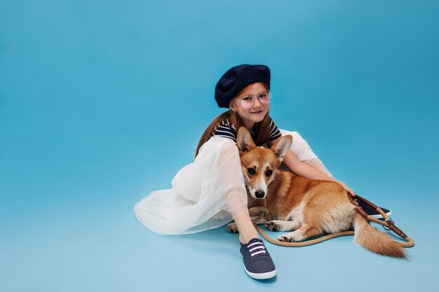 Vrij modieus jong meisje in moderne brillen blauwe baret sneakers en witte rok zittend op de vloer met haar kleine hond