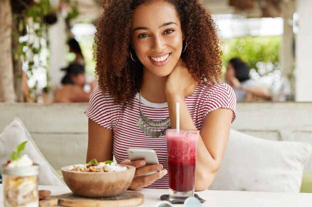 Vrij mooie jonge Afro-Amerikaanse vrouwelijke freelancer maakt instellingen op mobiele telefoon, drinkt smoothie en exotische zoete salade, besteedt vrije tijd op terras, heeft positieve uitdrukking en glimlach