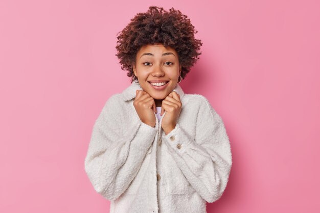 Vrij positieve jonge vrouw draagt kunstmatige bontjas glimlacht, voelt zich aangenaam blij poses tegen roze studio achtergrond. Portret van een vrouwelijk model met krullend haar voelt zich optimistisch in een goed humeur.
