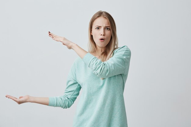 Vrij schattig blonde vrouw die vrijetijdskleding draagt die iets groots met handen laat zien, haar wenkbrauwen fronst, lippen pruilt.