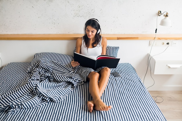 Vrij sexy lachende vrouw zittend in bed in de ochtend, luisteren naar muziek op de koptelefoon en leesboek