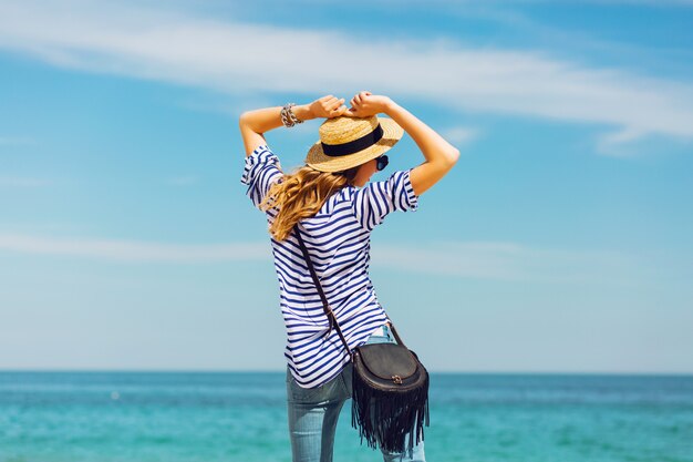 Vrij slanke bruine blonde stijlvolle vrouw in strooien hoed en zonnebril, die zich voordeed op het paradijs tropische strand