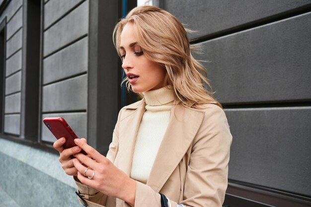 Vrij stijlvol blond meisje dat zorgvuldig mobiele telefoon gebruikt op straat in de stad