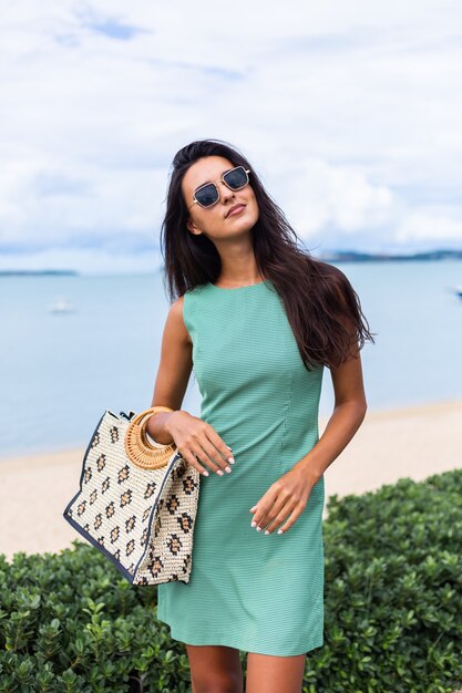 Vrij stijlvolle gelukkige vrouw in groene zomerjurk met tas, zonnebril dragen op vakantie, blauwe zee op achtergrond