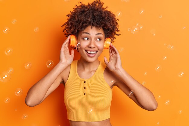 Vrij tevreden gekrulde harige vrouw draagt stereo koptelefoon op oren glimlacht in grote lijnen geniet van perfect geluid geconcentreerd weg gekleed in bijgesneden top geïsoleerd op oranje studio muur met vliegende zeepbellen