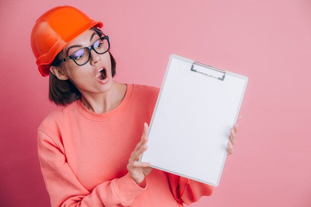 Vrij verbaasd verrast vrouw werknemer bouwer houdt wit bord leeg tegen roze achtergrond. Helm bouwen.