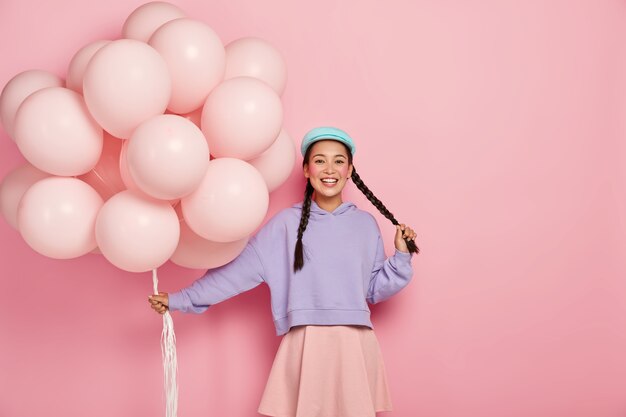 Vrij vrolijk Aziatisch tienermeisje komt op vakantie met een bos luchtballonnen, heeft twee donkere lange vlechten, rode wangen en minimale make-up, draagt een oversized paarse trui en rok, in een goed humeur