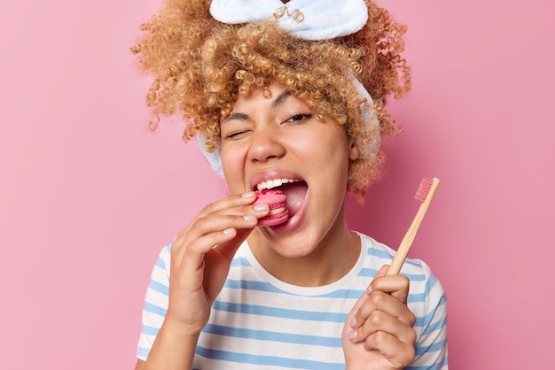 Vrij vrolijke vrouw eet heerlijke bitterkoekjes geeft niets om schadelijk voedsel voor je tanden houdt houten tandenborstel draagt gestreepte t-shirt poses binnen Schade van dessert op glazuur Cariës concept