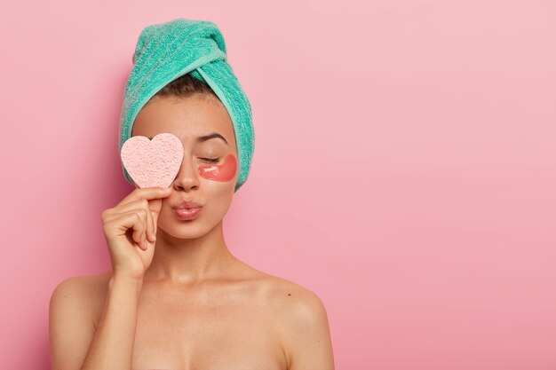 Vrij vrouwelijk model heeft gesloten ogen, afgeronde lippen, staat met blote schouders, geniet van schoonheidsbehandelingen, houdt cosmetische spons over het oog