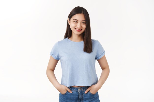 Vrolijk aangenaam vriendelijk assertief uitgaand aziatisch jong meisje hand in hand zakken jeans glimlachend breed wil helpen lichtblauw tshirt ontspannen zorgeloos pose staande witte achtergrond