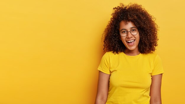 Vrolijk blij krullend schattig meisje lacht uit van geluk, verheugt zich op aangename momenten in het leven, heeft een aantrekkelijke uitstraling, draagt een grote transparante bril en een casual geel t-shirt, voelt zich vrolijk