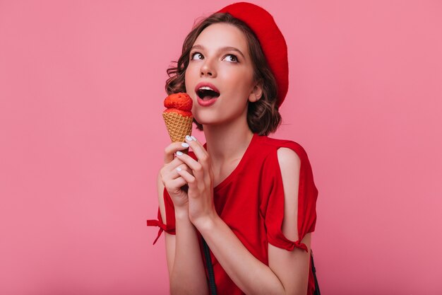 Vrolijk brunette meisje in het rode t-shirt poseren met ijs. Binnenfoto van innemende dame die in baret dessert eet.