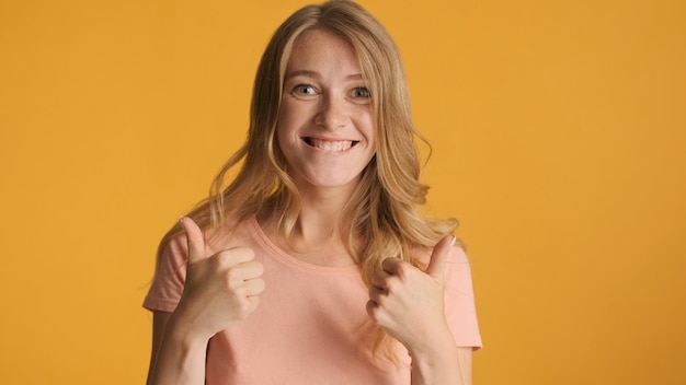 Vrolijk cool blond meisje dat duimen omhoog houdt en vreugdevol in de camera kijkt geïsoleerd op gele achtergrond