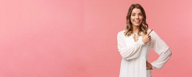 Vrolijk, goed uitziend Kaukasisch blond meisje in witte jurk wijzend naar de rechterbovenhoek en glimlachend naar ca