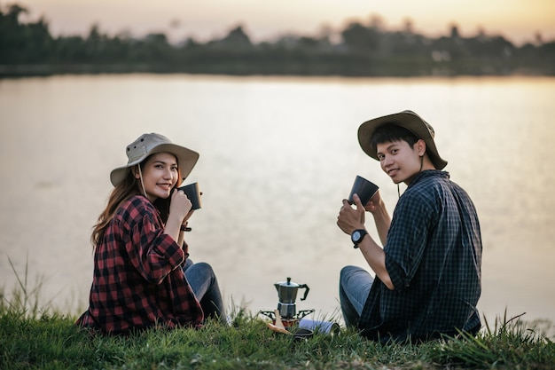 Vrolijk jong backpackerpaar zittend op het gras in de buurt van het meer in de vroege ochtend en het maken van verse koffiemolen tijdens het kamperen op zomervakantie