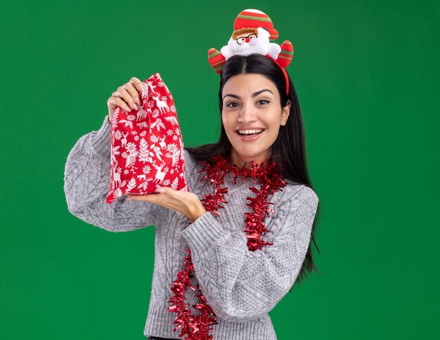 Vrolijk jong Kaukasisch meisje dat de hoofdband van de Kerstman en klatergoudslinger om hals draagt die de zak van de Kerstmisgift houdt die op groene muur met exemplaarruimte wordt geïsoleerd