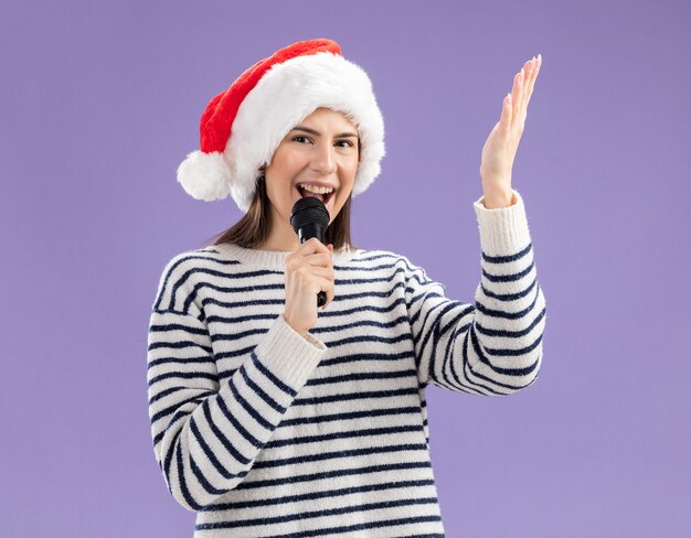 Vrolijk jong kaukasisch meisje met kerstmuts houdt microfoon vast die doet alsof ze zingt en houdt hand open geïsoleerd op paarse muur met kopieerruimte wall