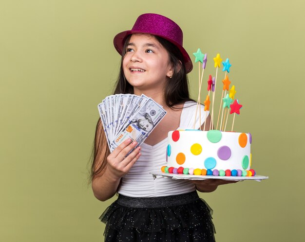 Vrolijk jong Kaukasisch meisje met paarse feestmuts met verjaardagstaart en geld kijkend naar kant geïsoleerd op olijfgroene muur met kopieerruimte