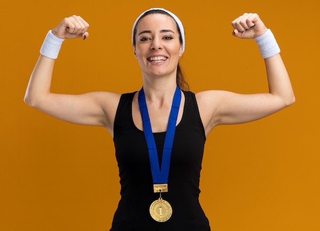 Gratis foto vrolijk jong, vrij sportief meisje met een hoofdband en polsbandjes met een medaille om de nek en doet een sterk gebaar geïsoleerd op een oranje muur