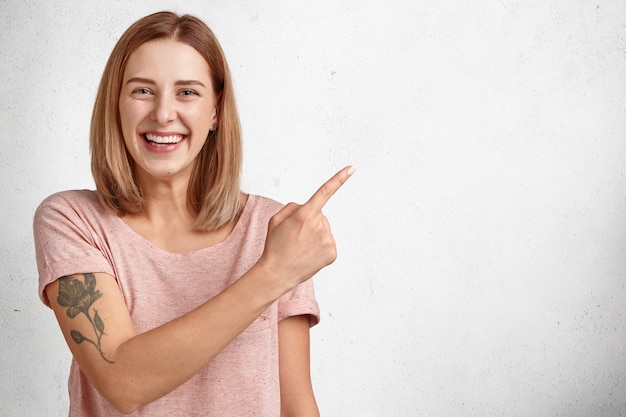 Vrolijk jong vrouwenmodel met kort kapsel, brede glimlach, wijst op rechtsboven, gekleed in vrijetijdskleding