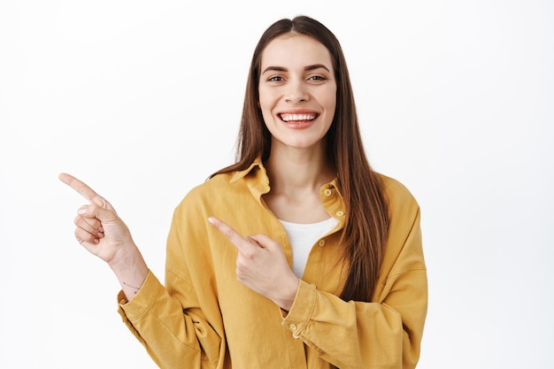 Vrolijk Kaukasisch vrouwelijk model glimlachend en wijzend met de vingers opzij, uitnodigend om de winkelwebsite te bezoeken, advertenties op kopieerruimte te tonen, informatie weer te geven, staande op een witte achtergrond