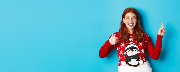Vrolijk kerstfeest vrolijk roodharig meisje in kerstsweater wijzende vinger naar rechterbovenhoek tonen