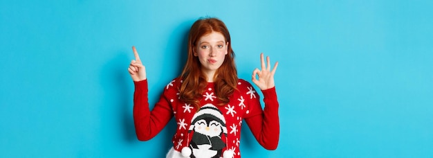 Gratis foto vrolijk kerstfeest vrolijk roodharig meisje in kerstsweater wijzende vinger naar rechterbovenhoek tonen