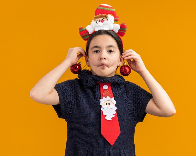 Vrolijk meisje in gebreide jurk met rode stropdas met grappige rand op het hoofd met kerstballen over haar oren gelukkig en positief grimas makend over oranje muur