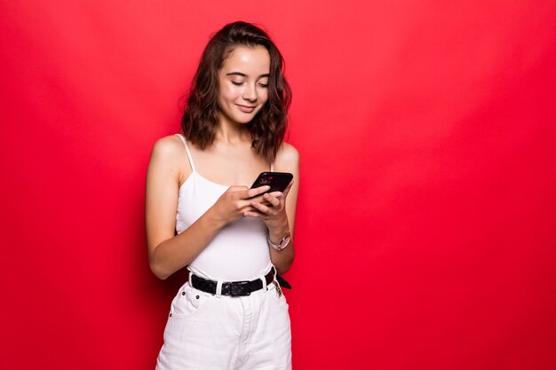 Vrolijk meisje typt iets op haar mobiele telefoon geïsoleerd op rode muur