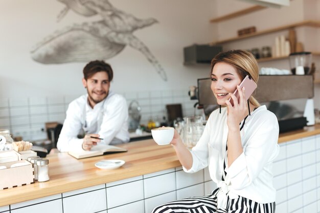 Vrolijk meisje zit aan de balie met een kopje koffie in de hand en praat graag op haar mobiele telefoon terwijl de barista naar haar kijkt in de coffeeshop Mooie dame met blond haar die koffie drinkt in café