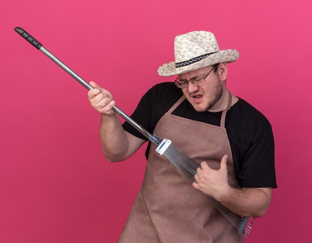 Vrolijk met gesloten ogen jonge mannelijke tuinman met een tuinhoed met bladhark zingend geïsoleerd op roze muur