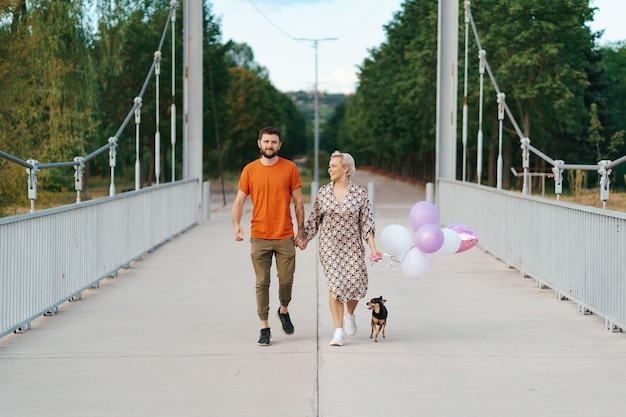 Vrolijk mooi paar die gelukkig op brug met hun hond en roze ballons glimlachen lopen