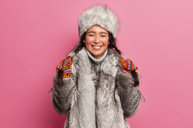 Vrolijk noordelijk meisje houdt twee vlechten in handen glimlacht in grote lijnen draagt winterkleren bereidt zich voor op uitgaan tijdens koude dag vormt tegen roze studiomuur