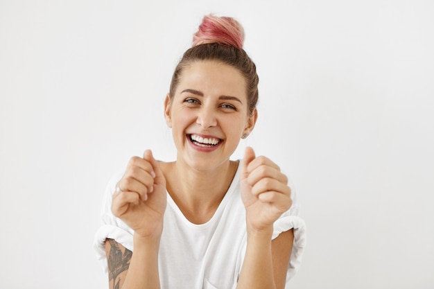 vrolijk opgewonden jonge dame met getatoeëerde armen en pastelroze haarknoop die haar succes en prestatie op het werk viert, poseert met gebalde vuisten, haar ogen die opwinding uitdrukken