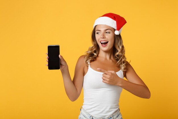 Vrolijk santa meisje in kerstmuts poseren geïsoleerd op gele achtergrond. gelukkig nieuwjaar 2020 viering vakantie concept. bespotten kopie ruimte. wijzende vinger op mobiele telefoon met leeg leeg scherm.