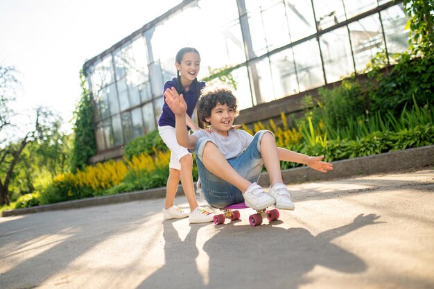 Vrolijk schattig donkerharig meisje dat een vriend helpt om zittend op het skateboard te rijden