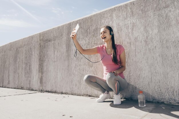 Vrolijk sportmeisje dat buiten zit, naar muziek luistert op een koptelefoon en in haar smartphone kijkt