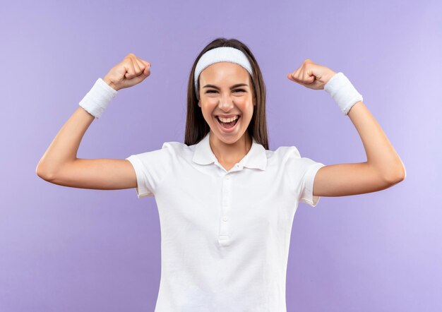 Vrolijk vrij sportief meisje met hoofdband en polsbandje gebaren sterk geïsoleerd op paarse muur purple