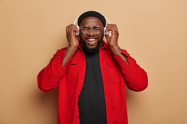 Vrolijk zwart ongeschoren mannelijk model verheugt zich op het luisteren naar favoriete muziek in de headset, loenst gezicht van gelach