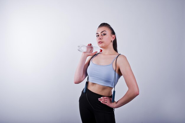 Vrolijke aantrekkelijke jonge fitness vrouw in top en zwarte legging met springtouw en fles water geïsoleerd op witte achtergrond