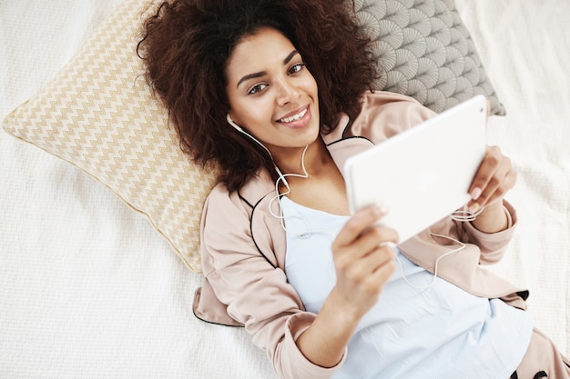 Vrolijke Afrikaanse vrouw in nachtkleding en hoofdtelefoons die holdingstablet glimlachen die op bed liggen.