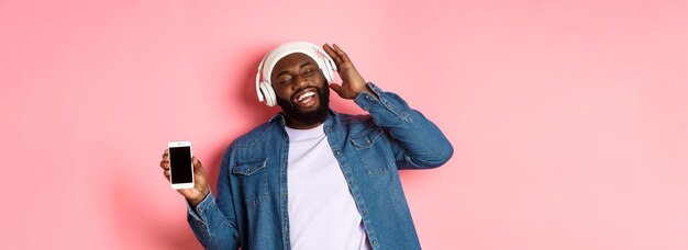 Vrolijke Afro-Amerikaanse hipster-kerel die muziek luistert in een koptelefoon die de app voor het telefoonscherm laat zien en zingt