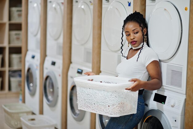 Vrolijke afro-amerikaanse vrouw met witte mand in de buurt van wasmachine in de zelfbedieningswasserij