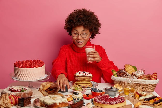 Vrolijke Afro-vrouw strekt zich uit naar een heerlijk dessert, houdt een glas melk vast, eet cake, omringd met junkfood, draagt een bril en een rode trui, kan geen nee zeggen tegen snoep