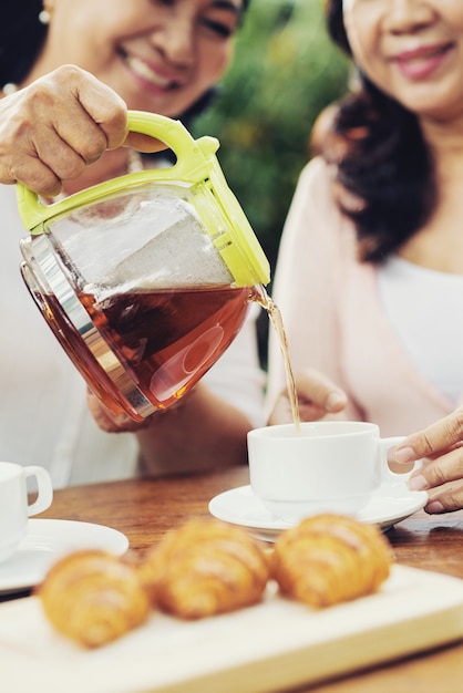 Vrolijke aziatische dames die thee van theepot in koppen gieten en croissants op tafel