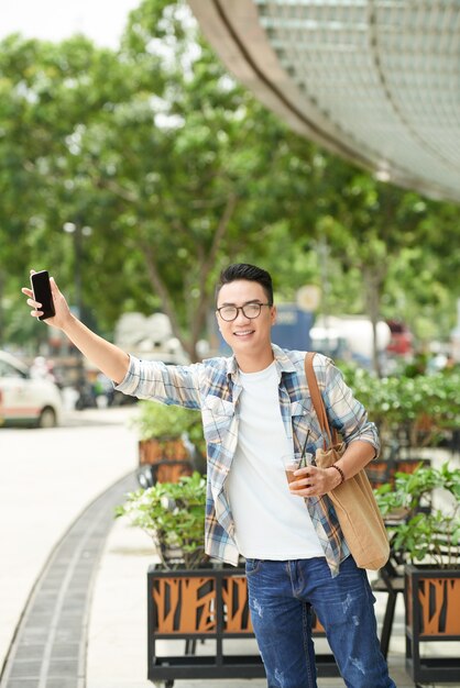 Vrolijke Aziatische mens die zich naast weg bevindt en hand met smartphone uitsteekt