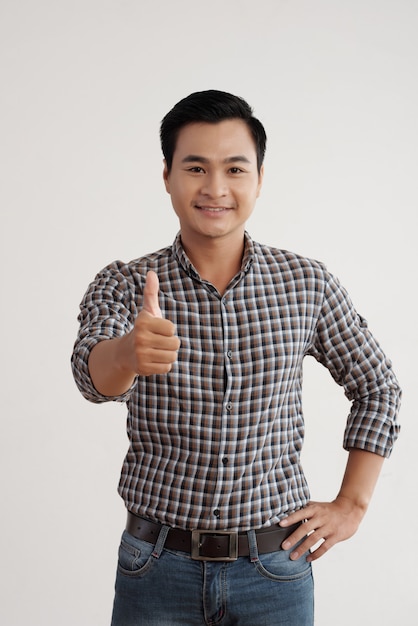 Vrolijke Aziatische mens in plaidoverhemd en jeans die zich in studio met zijn omhoog duim bevinden