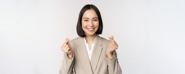Vrolijke Aziatische verkoopster glimlachend en vinger harten teken staande in pak op witte achtergrond tonen